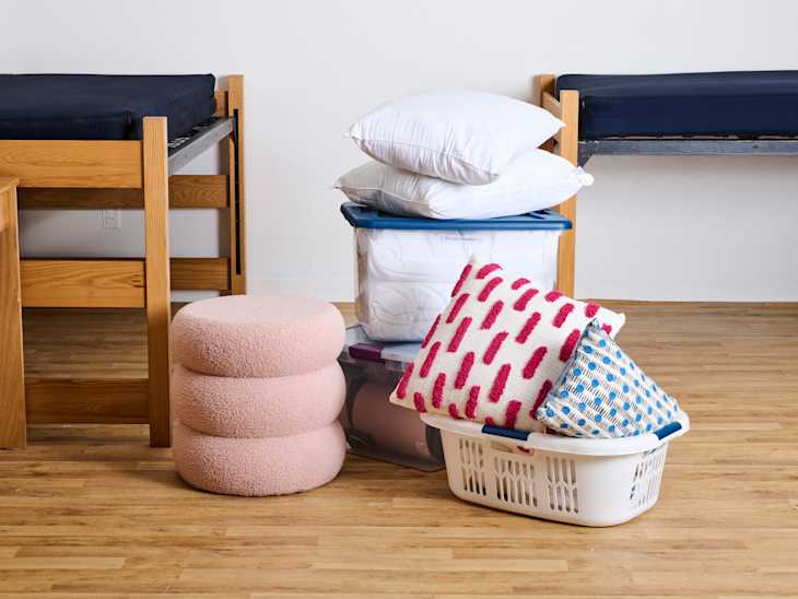 A stack of pillows and colorful cushions next to a laundry basket in a dorm room with wooden furniture.