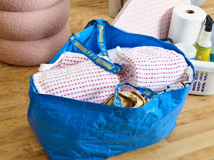 A blue tote bag filled with patterned towels, cleaning supplies, and a white basket on a wooden floor.