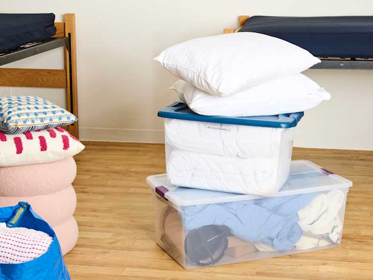 Bedding and storage bins stacked in a room with wooden floors and two beds in the background.