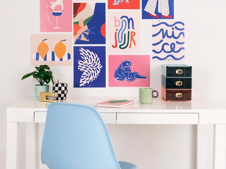 A modern desk with a light blue chair, colorful wall art, a small plant, and stacked storage boxes.