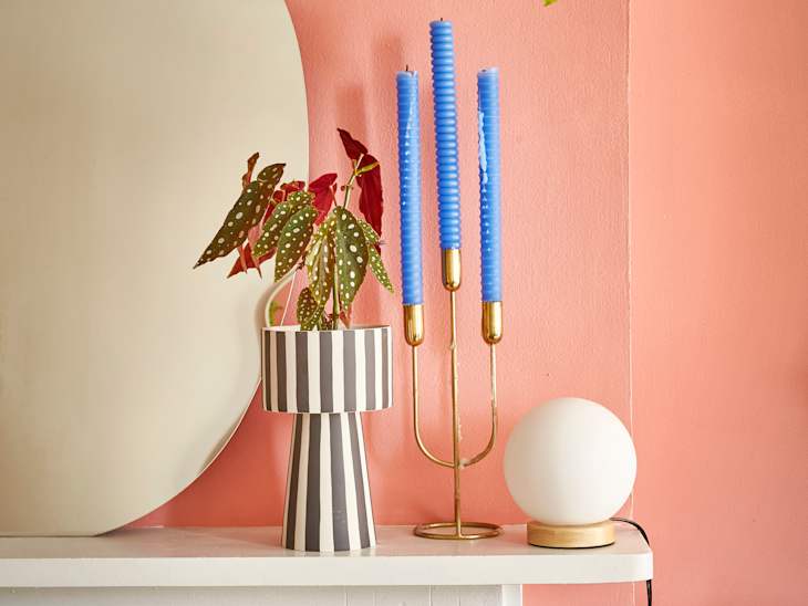 A striped vase with a begonia plant, blue spiral candles in a gold holder, and a round white lamp on a pink wall.