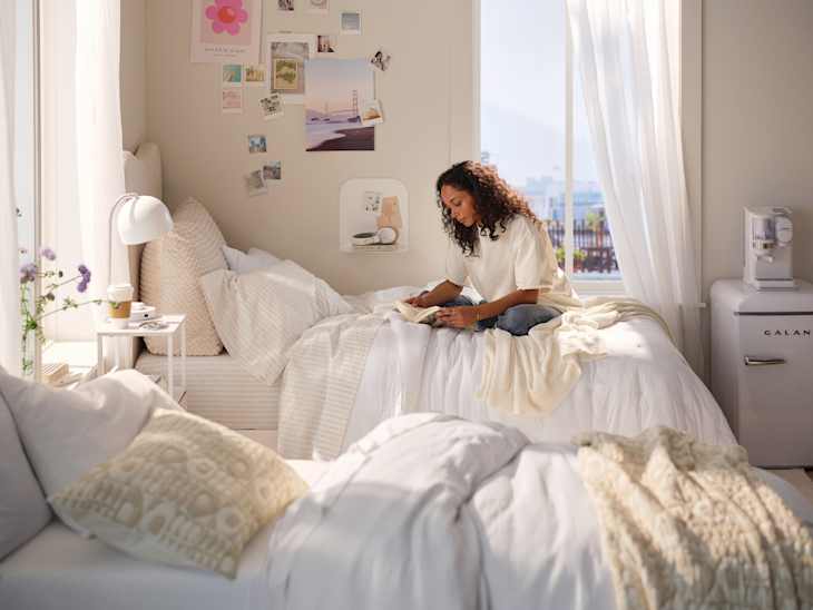 A cozy bedroom with two beds, a woman reading on one, soft blankets, a small table, and a mini fridge.
