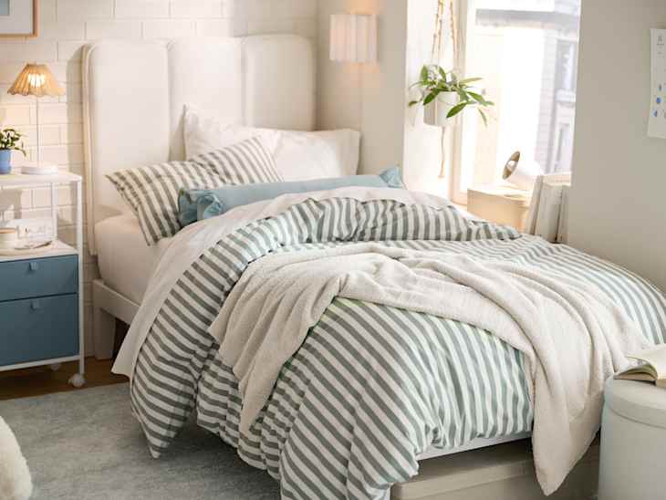 Cozy bedroom with a striped duvet, white headboard, bedside table, lamp, and a plant by the window.