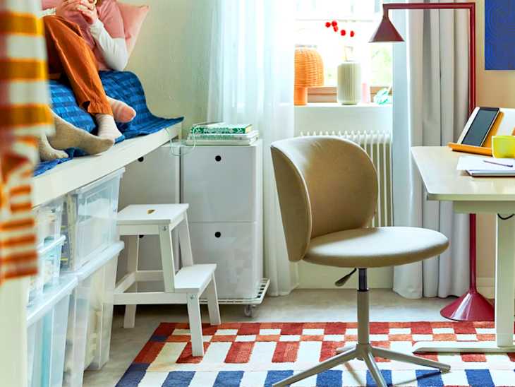 Cozy bedroom corner with a blue bedspread, a desk with a tablet, a beige swivel chair, and colorful rug.