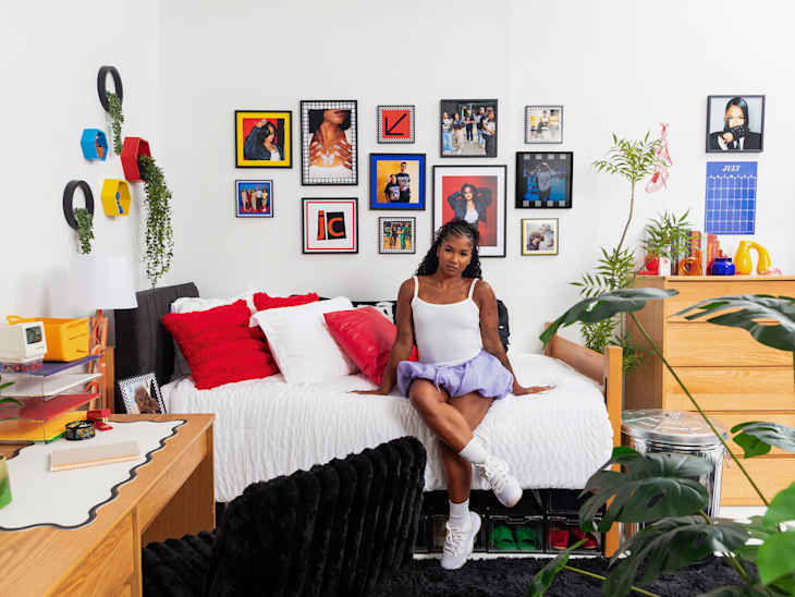 A young woman sitting on a bed with red and white pillows in a colorful bedroom featuring framed photos, plants, and a desk.