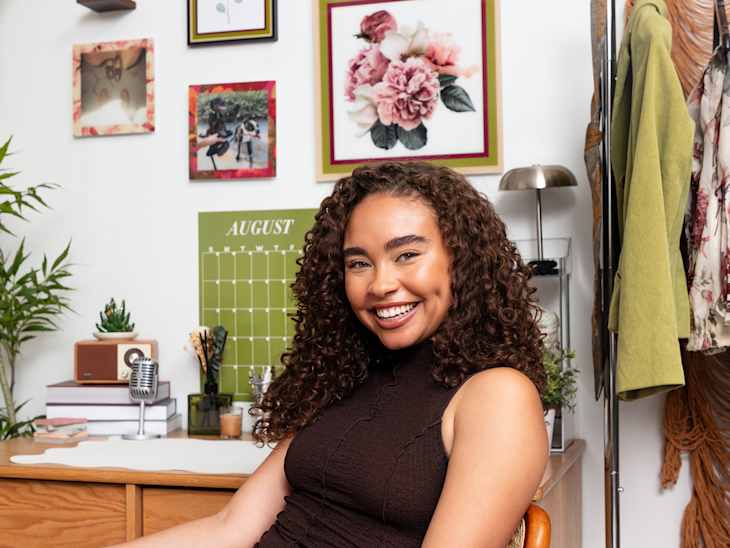 A young woman with curly hair smiles while sitting in a chair, surrounded by a colorful gallery wall and a desk with decor items.