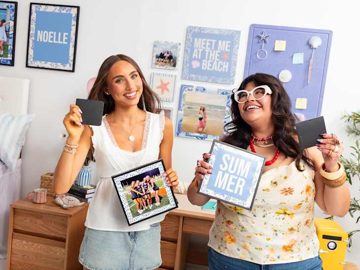Two women smiling and holding photo frames in a colorful, decorated room with beach-themed wall art.