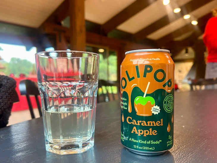 A glass of clear liquid next to a can of OLIPOP Caramel Apple soda on a dark table in a rustic setting.