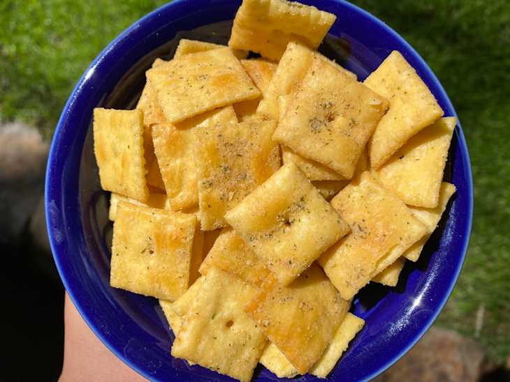Bowl filled with seasoned square crackers, held against a green grass background.
