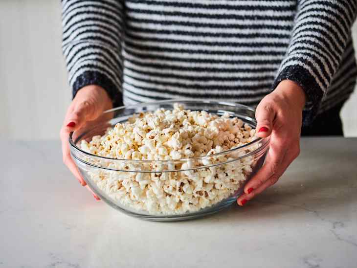 Person holding a glass bowl filled with freshly popped popcorn on a marble countertop.