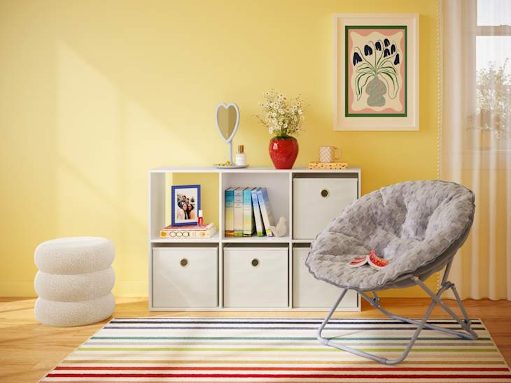 Cozy room with yellow walls featuring a white shelf, a gray papasan chair, and a striped rug. Decor includes books, a photo frame, and a vase.