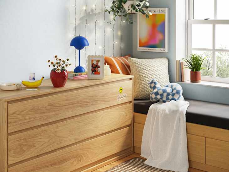 Cozy bedroom corner with a wooden dresser, blue lamp, potted plants, and decorative pillows on a window seat.