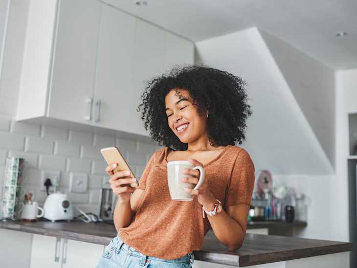 Shot of a young woman using a smartphone and having coffee in the kitchen at home.