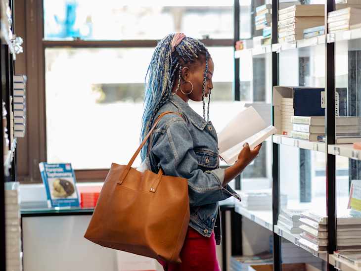 A black female student in her 20's standing looking through books in the library.
