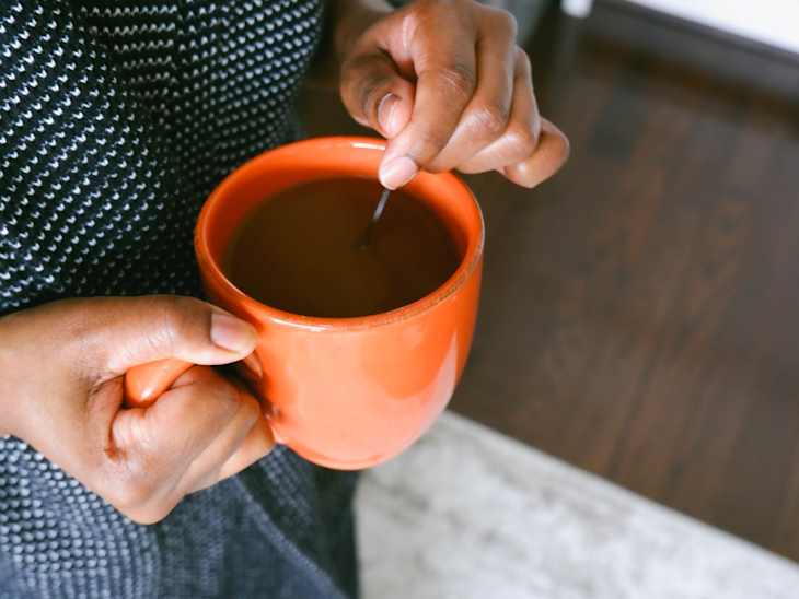 view of woman's hands stirring coffee in an orange mug. rug and wood floor visible below.