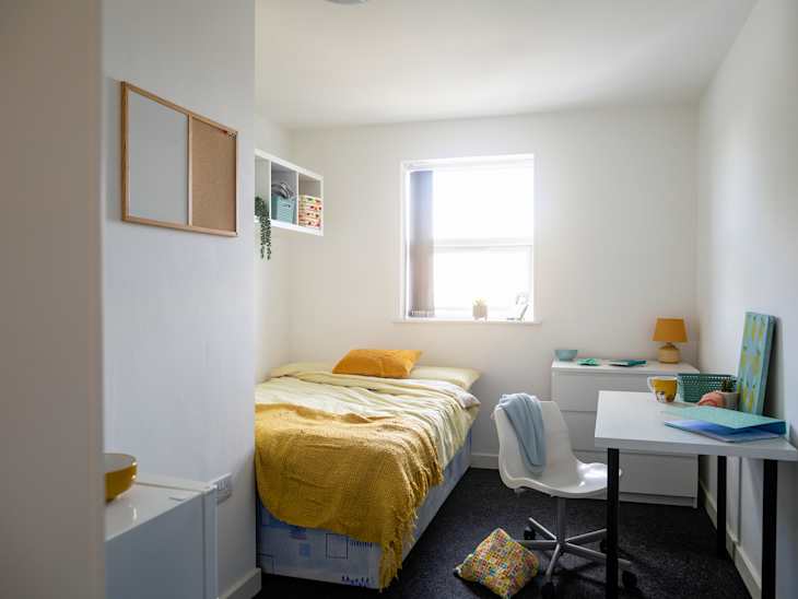 Cozy bedroom with a bed covered in yellow and green bedding, a desk with a chair, and shelves displaying decor items.