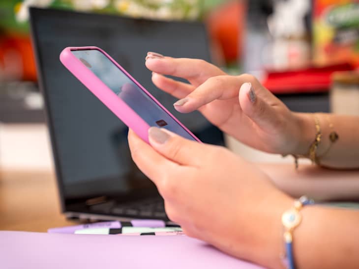Smartphone in female hands. Close up of hands with a phone. In the background there is a laptop on the desk. Indoor photo