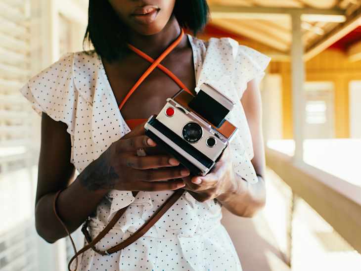 woman wearing white dress with small black polka dots holding white polaroid camera on a brown leather strap