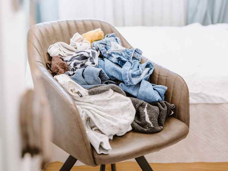 Pile of unfolded dirty clothes for laundry on the chair. Heap of used clothes for donation and recycling. Concept of minimalism, mess and wardrobe cleaning