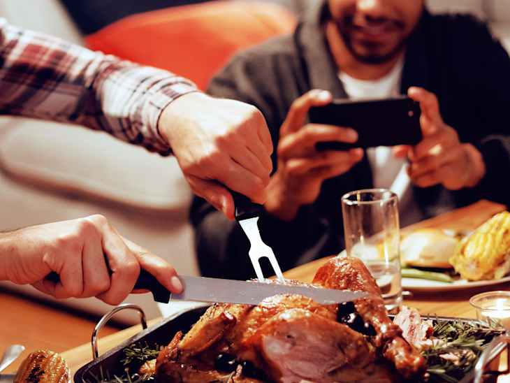 Front view of a young mixed race man sitting at a table for Thanksgiving dinner at home with friends carving the turkey, a young mixed race male friend taking a photo with his smartphone in the