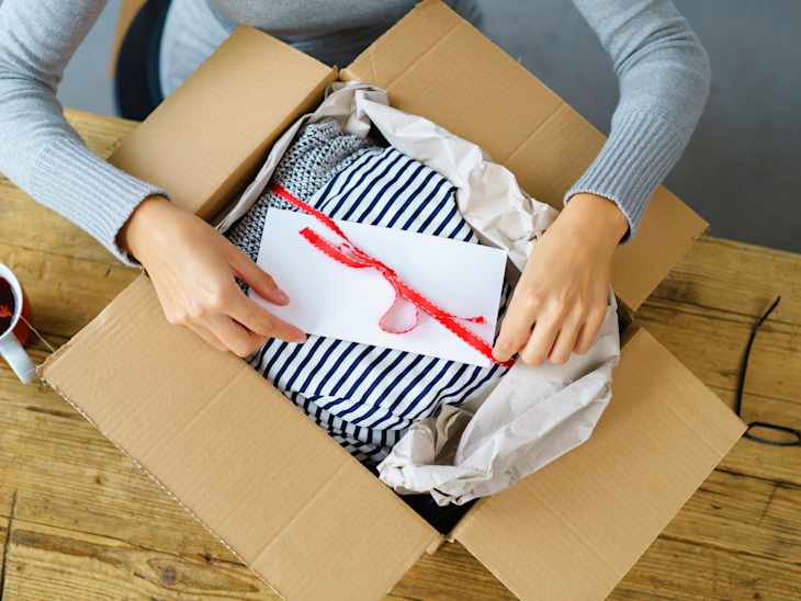 Person opening a cardboard box with a striped gift wrapped in tissue paper, next to a cup of tea on a wooden table.