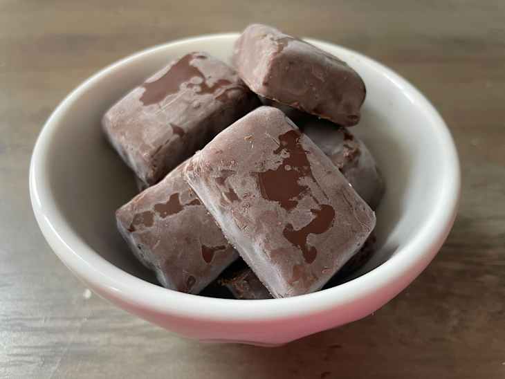 Frozen chocolate-covered treats stacked in a white bowl on a wooden surface.