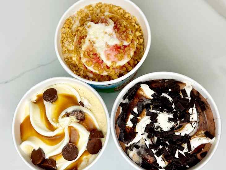 Three cups of ice cream: one with crumbled topping and strawberry sauce, one with caramel and chocolate pieces, and one with chocolate swirls and shavings.