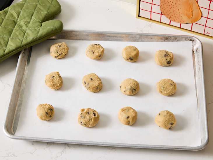 Cookie dough balls with chocolate chips arranged on a baking sheet lined with parchment paper, green oven mitt nearby.