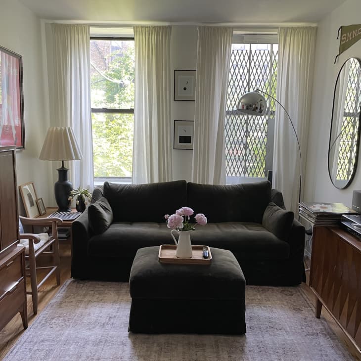 Living room with dark sofa, ottoman, wooden furniture, floor lamp, and wall-mounted TV, featuring large windows with curtains.