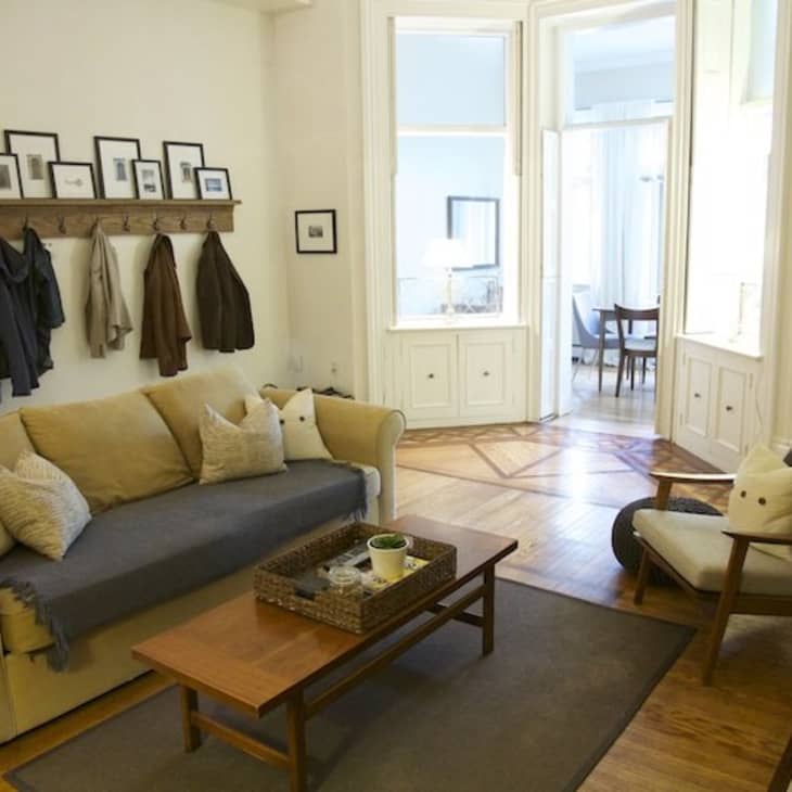 Living room with beige sofa, wooden coffee table, framed photos, and coats on hooks, leading to a dining area.