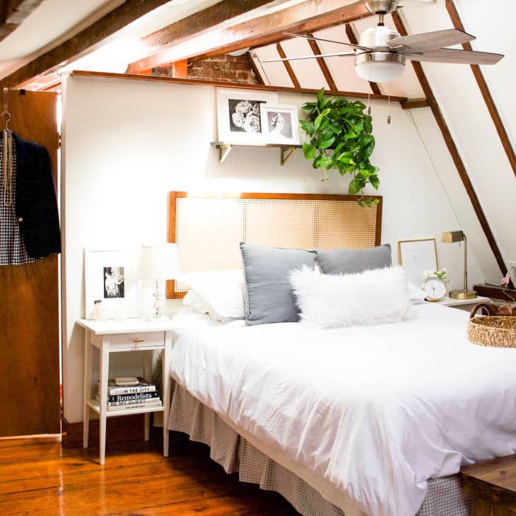 Cozy attic bedroom with a white bed, wicker headboard, hanging plant, and wooden beams.