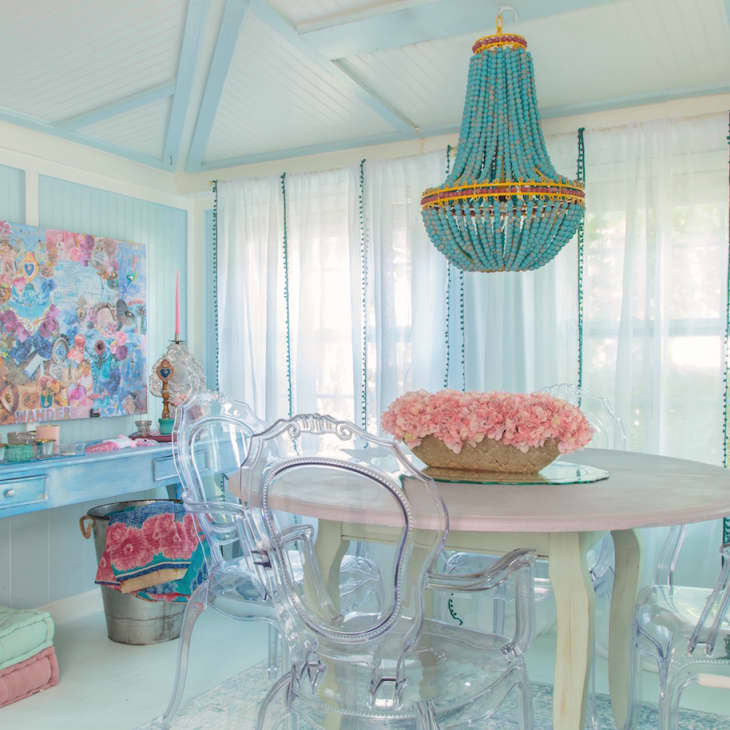 Dining room with a round table, pink flowers, clear chairs, blue chandelier, and a colorful art piece on a blue desk.