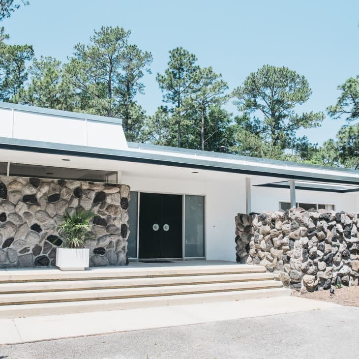 Mid-century modern house with stone facade, large windows, and surrounded by pine trees.