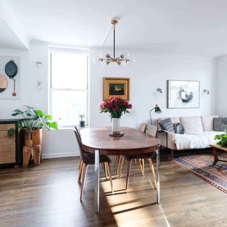 Open-plan living and dining area with wooden table, beige sofa, plants, and wall art, featuring a modern chandelier.