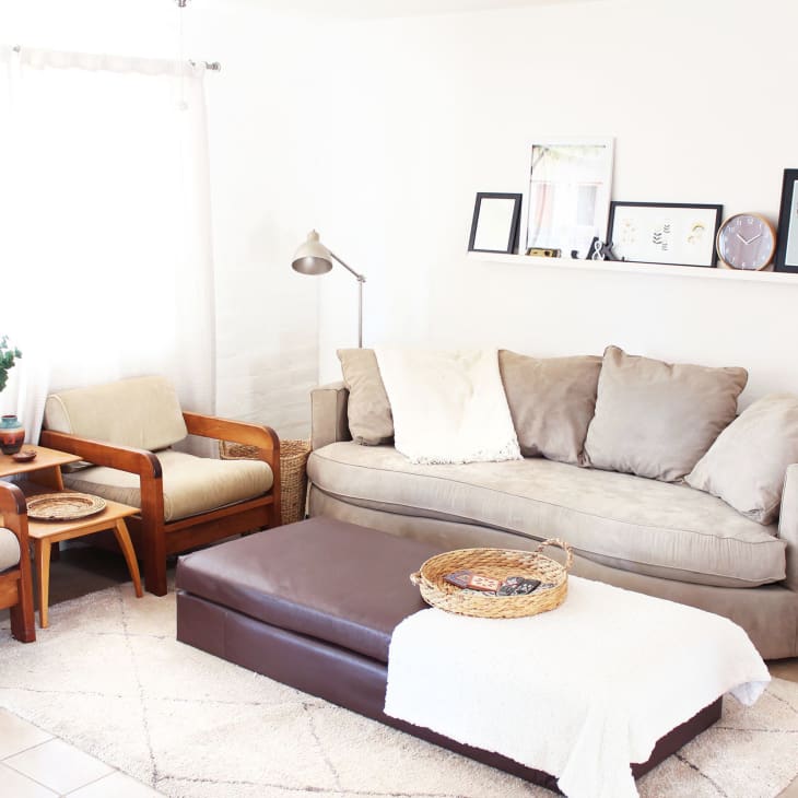 Living room with beige sofa, two wooden armchairs, a brown ottoman, and wall shelf with framed art and a clock.