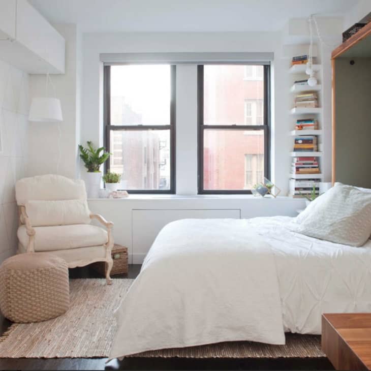 Cozy bedroom with a white bed, armchair, woven ottoman, bookshelves, and large window.
