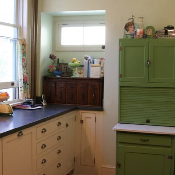 Vintage kitchen with cream cabinets, black countertop, green cupboard, and floral curtains near a window.