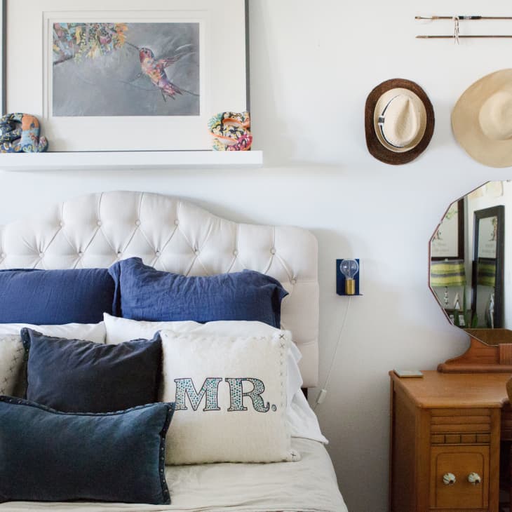 Tufted headboard with blue and white pillows, framed bird art, hats on wall, and wooden vanity with mirror.