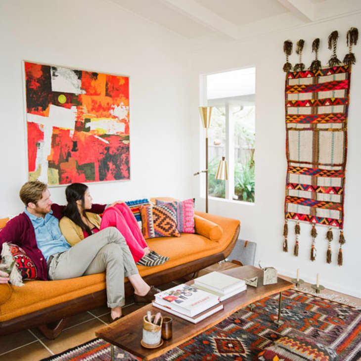 Couple sitting on an orange sofa in a living room with colorful abstract art, patterned pillows, and a woven wall hanging.