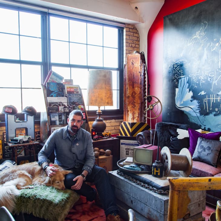 Man sitting with a dog in an eclectic room with vintage decor, large windows, and a chalkboard wall.
