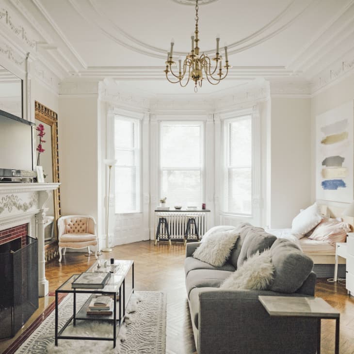 Elegant living room with ornate fireplace, gray sofa, glass coffee table, chandelier, and large bay windows.