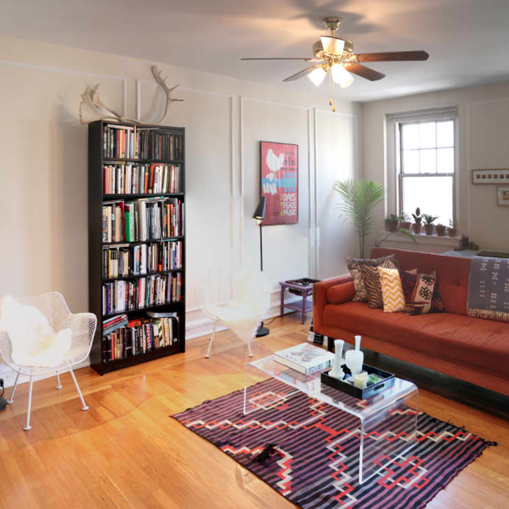 Living room with a rust-colored sofa, patterned pillows, black bookshelf, glass coffee table, and geometric rug.