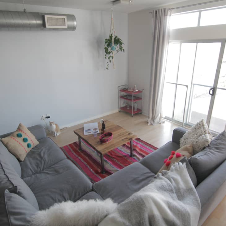 Gray sectional sofa with colorful pillows, wooden coffee table on red striped rug, and large windows in a modern living room.