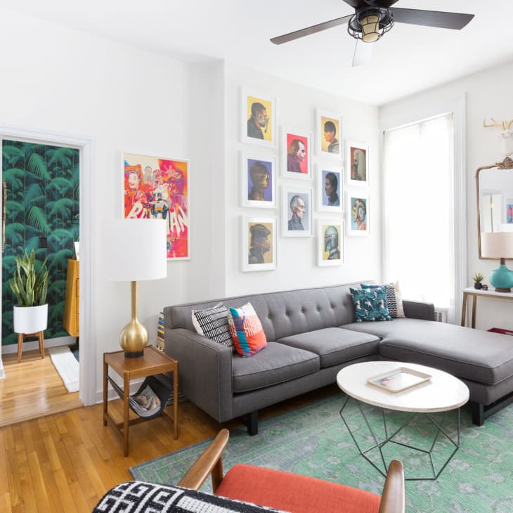 Living room with gray sectional sofa, colorful art gallery wall, round coffee table, and green rug. Doorway leads to plant-filled room.