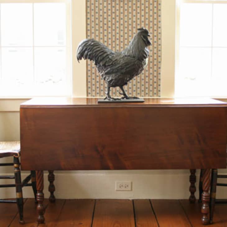 Wooden drop-leaf table with a rooster sculpture, flanked by two chairs in a sunlit room.