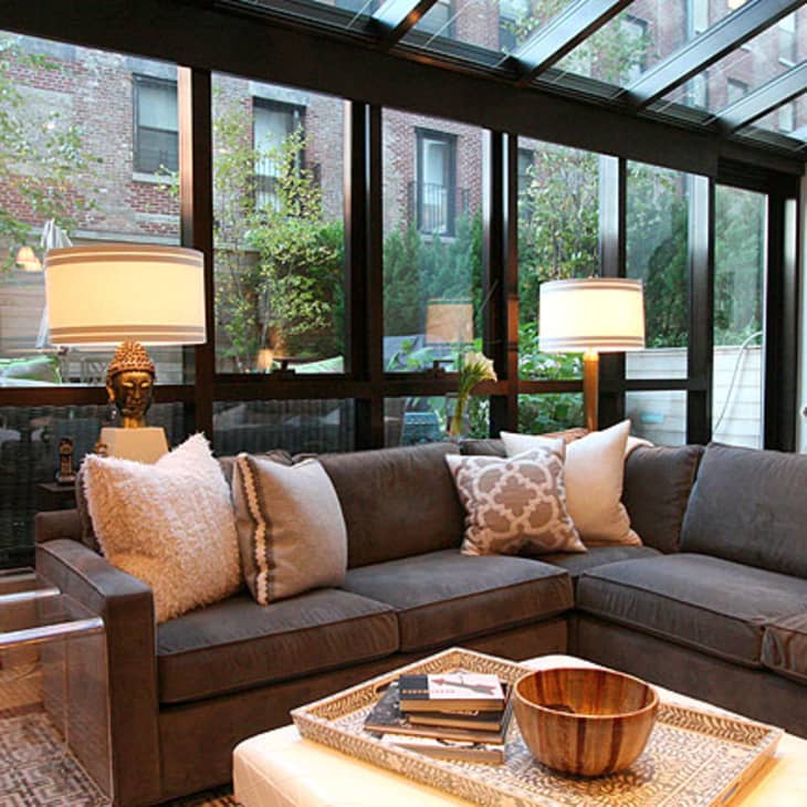 Sunroom with gray sectional sofa, decorative pillows, glass ceiling, and large windows overlooking a garden.