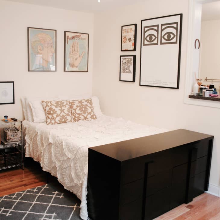 Bedroom with white bedding, patterned pillows, wall art, and a black dresser. Small kitchen area visible in the background.