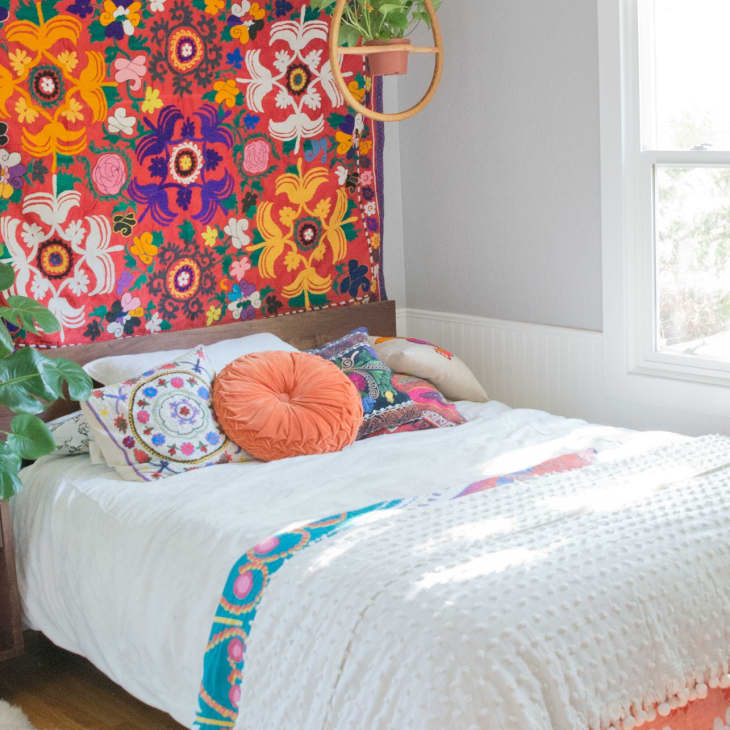 Bohemian bedroom with colorful floral tapestry, white bedding, and vibrant pillows, next to a window and green plants.