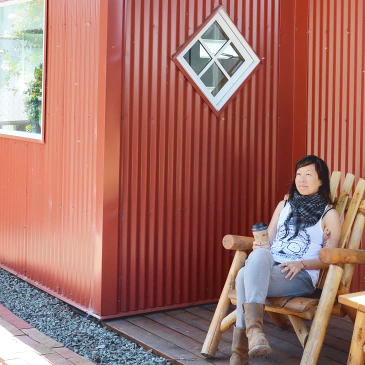 Woman sitting on a wooden chair outside a red corrugated building, holding a coffee cup, with potted plants on a table.