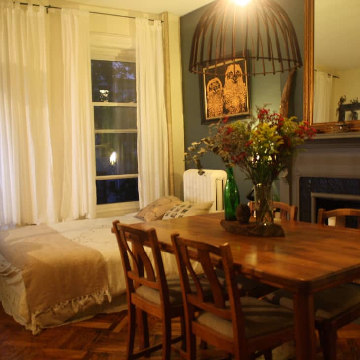 Cozy dining room with wooden table, chairs, floral centerpiece, large mirror, and bed by curtained windows.